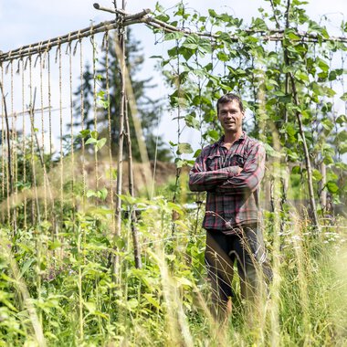 A man stands smiling in a garden with tall plants. In the background, there are wooden trellises and a green landscape. | © Kleinwalsertal Tourismus | Dominik Berchtold