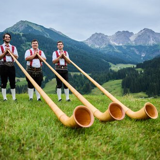 © Kleinwalsertal Tourismus eGen | Oliver Farys Drei Männer in traditioneller Tracht stehen in einer grünen Wiese mit einer beeindruckenden Berglandschaft im Hintergrund. Sie halten große Alphörner in den Händen. | © Kleinwalsertal Tourismus eGen | Oliver Farys