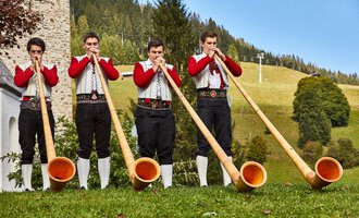 Vier junge Männer in traditioneller Kleidung spielen auf Alphörnern in einer malerischen Landschaft. Im Hintergrund sind grüne Wiesen und Bäume zu sehen. | © Kleinwalsertal Tourismus | Frank Drechsel