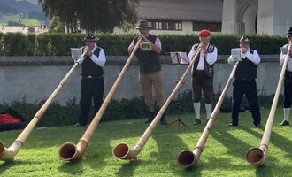 A group of five men is playing alphorns on a meadow. They are wearing traditional costumes and are standing in a picturesque setting. | © Alphornfreunde grenzenlos
