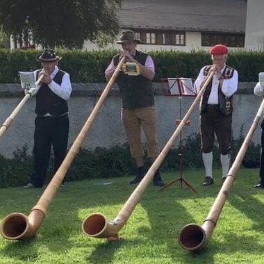 A group of five men is playing alphorns on a meadow. They are wearing traditional costumes and are standing in a picturesque setting. | © Alphornfreunde grenzenlos