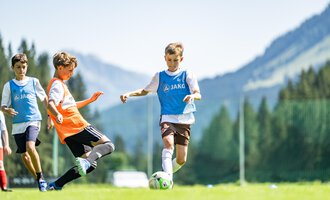 Junge Fußballspieler auf einem grünen Spielfeld mit Bergen im Hintergrund. Ein Spieler dribbelt den Ball, während ein anderer ihn versucht zu stoppen. | © Fußballschule Anstoß