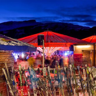Eine Skihütte mit roten Sonnenschirmen in einem winterlichen Ambiente. Im Vordergrund stehen mehrere Skier und im Hintergrund sind Menschen zu sehen, die die Atmosphäre genießen. | © Plattform Kleinwalsertal | Michael Monschau