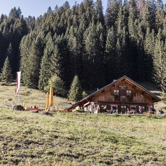© Kleinwalsertal Tourismus | Bastian Morell Eine rustikale Hütte inmitten von grünen Wiesen und dichten Nadelbäumen. Der Himmel ist klar und die Umgebung wirkt einladend und friedlich. | © Kleinwalsertal Tourismus | Bastian Morell