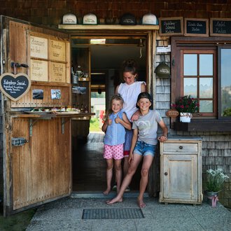 © Kleinwalsertal Tourismus | Oliver Farys Eine einladende Eingangstür zu einem kleinen Laden, umgeben von Holz und Grün. Drei Kinder stehen fröhlich davor und lächeln in die Kamera. | © Kleinwalsertal Tourismus | Oliver Farys