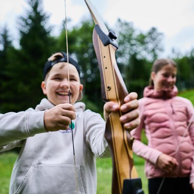 Zwei Personen im Wald beim Bogenschießen. Die Frau trägt einen bunten Pullover und zielt mit einem Bogen, während der Mann in einem grünen T-Shirt hinter ihr steht. | © Kleinwalsertal Tourismus eGen - Fotograf: Oliver Farys