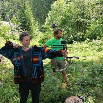 © Wildnisschule Kleinwalsertal | Ronny Katz Zwei Personen im Wald beim Bogenschießen. Die Frau trägt einen bunten Pullover und zielt mit einem Bogen, während der Mann in einem grünen T-Shirt hinter ihr steht. | © Wildnisschule Kleinwalsertal | Ronny Katz