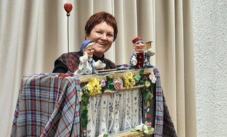 © Marie Luise Kaiser | Veronika Senn A woman presents a puppet show with two handcrafted puppets. The background is adorned with a bright curtain and colorful decorations. | © Marie Luise Kaiser | Veronika Senn