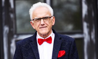 An older man with gray hair is wearing an elegant suit and a red bow tie. He is holding a piece of paper in his hand and standing in front of a building with large windows. | © Johann Mösenbichler - Fotograf: Tobias Epp