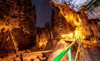 A narrow, snow-covered path winds through a deeply snow-laden gorge. The night sky is dotted with stars, while warm lights illuminate the surroundings. | © Breitachklammverein eG | Dominik Berchtold