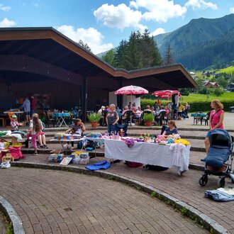 © @Flohmarkt im Kurpark Riezlern Kulturcafe A lively outdoor flea market with many tables full of goods. In the background, there are stalls, children, and a picturesque mountain landscape. | © @Flohmarkt im Kurpark Riezlern Kulturcafe