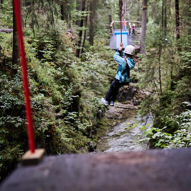 Ein Kind schwingt an einer Seilrutsche zwischen Bäumen in einem grünen Wald. Umgeben von Natur und einem schmalen Bach darunter. | © Kleinwalsertal Tourismus | Oliver Farys