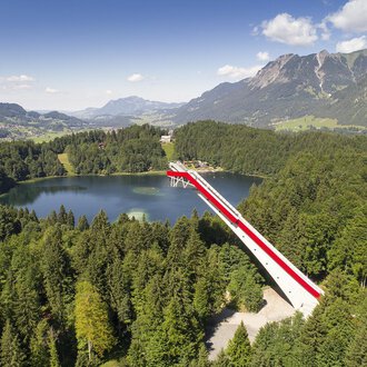 © Skiflugschanze Oberstdorf | Eren Karaman Eine beeindruckende Aussichtsbrücke über einen klaren See, umgeben von grünen Wäldern und Bergen. Der Himmel ist blau mit einigen Wolken. | © Skiflugschanze Oberstdorf | Eren Karaman