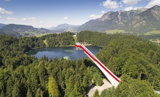 Eine beeindruckende Aussichtsbrücke über einen klaren See, umgeben von grünen Wäldern und Bergen. Der Himmel ist blau mit einigen Wolken. | © Skiflugschanze Oberstdorf | Eren Karaman