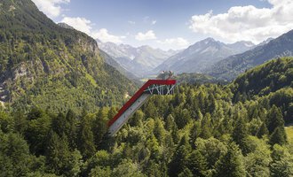 Eine beeindruckende Aussichtsplattform, die über die Bäume ragt, umgeben von majestätischen Bergen und dichten Wäldern. Der Himmel ist klar und zeigt einige Wolken. | © Skiflugschanze Oberstdorf | Eren Karaman