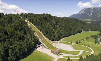Eine beeindruckende Skisprungschanze umgeben von grünen Wäldern und Bergen. Die Landschaft ist malerisch und zeigt eine Mischung aus Natur und Sporteinrichtungen. | © Skiflugschanze Oberstdorf | Eren Karaman