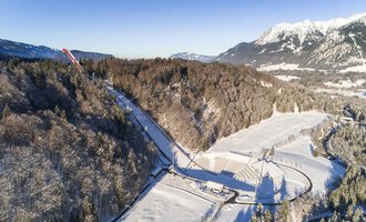 Eine schneebedeckte Landschaft mit Bäumen und einem klaren Himmel. Im Hintergrund sind Berge zu sehen, die die winterliche Szenerie ergänzen. | © Skiflugschanze Oberstdorf | Eren Karaman