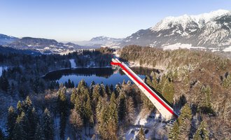 Eine lange Brücke mit roten und weißen Streifen führt über ein ruhiges, schneebedecktes Tal und einen See. Im Hintergrund sind majestätische Berge zu sehen, die in der Wintersonne erstrahlen. | © Skiflugschanze Oberstdorf | Eren Karaman