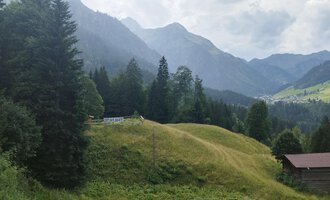 Eine malerische Landschaft mit grünen Hügeln und umgeben von hohen Bergen. Im Vordergrund steht eine kleine Hütte, umgeben von Bäumen. | © Nicole Lughammer
