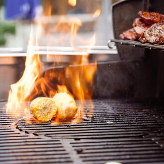 © Hotel Auenhütte | Eva Backes Ein Grill mit Flammen, auf dem Lebensmittel gekocht werden. Man sieht gegrillte Kartoffeln und Fleisch. | © Hotel Auenhütte | Eva Backes