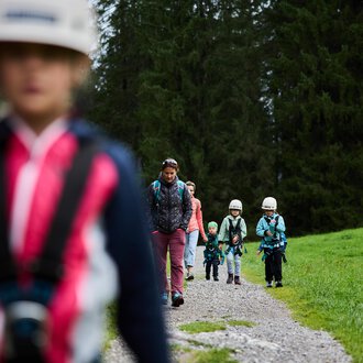 © Kleinwalsertal Tourismus | Oliver Farys Eine Gruppe von Kindern wandert auf einem Schotterweg durch einen Wald. Sie tragen Helme und sind gut ausgestattet für ein Abenteuer in der Natur. | © Kleinwalsertal Tourismus | Oliver Farys