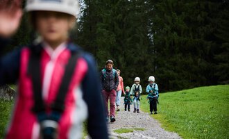 Eine Gruppe von Kindern wandert auf einem Schotterweg durch einen Wald. Sie tragen Helme und sind gut ausgestattet für ein Abenteuer in der Natur. | © Kleinwalsertal Tourismus | Oliver Farys