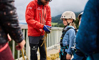 Ein Trainer erklärt einem Kind die sichere Handhabung von Kletterausrüstung. Im Hintergrund sind weitere Kinder und eine grüne Berglandschaft zu sehen. | © Kleinwalsertal Tourismus | Oliver Farys