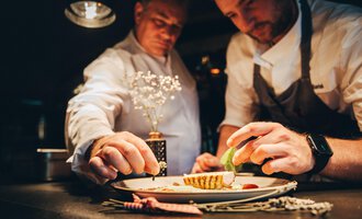 Two chefs working on a plate while carefully adding ingredients. The atmosphere is warm and inviting, with a focus on the artistic presentation of the dish. | © Hotel Birkenhöhe Bantel GmbH & Co KG