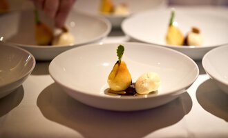 A beautifully arranged plate with an upright pear, served with a scoop of ice cream and a green decoration. The background shows several similar plates. | © Hotel Birkenhöhe | Annette Sandner