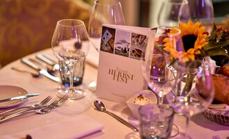 An elegantly set table with glasses and cutlery. In the middle, there is a menu card and a flower arrangement with sunflowers. | © Hotel Birkenhöhe | Annette Sandner