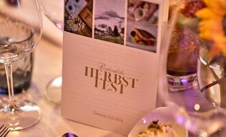 An elegantly set table with glasses and a menu for the autumn festival. In the background, there are flowers and a small bowl with a delicate dish. | © Hotel Birkenhöhe | Annette Sandner