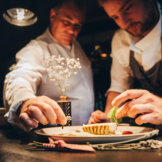 Zwei Köche, die an einem Teller arbeiten, während sie sorgfältig Zutaten hinzufügen. Die Atmosphäre ist warm und einladend, mit einem Fokus auf das kunstvolle Anrichten des Gerichts. | © Hotel Birkenhöhe Bantel GmbH & Co KG