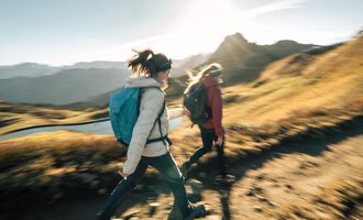 Zwei Wanderer gehen einen Pfad in den Bergen entlang. Die Sonne scheint und die Landschaft ist grün und goldfarben. | © Kleinwalsertal Tourismus eGen - Fotograf: Hannah Bichay