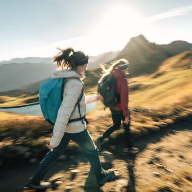 Zwei Wanderer gehen einen Pfad in den Bergen entlang. Die Sonne scheint und die Landschaft ist grün und goldfarben. | © Kleinwalsertal Tourismus eGen - Fotograf: Hannah Bichay