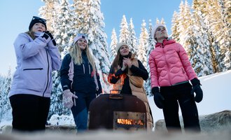 Eine Gruppe von Frauen steht um ein Feuer in einer verschneiten Landschaft. Im Hintergrund sind hohe, schneebedeckte Bäume und ein klarer blauer Himmel zu sehen. | © Kleinwalsertal Tourismus | Oliver Farys