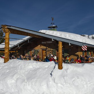 © Bergstüble | Michaela Beck Dornach A cozy mountain cabin in the snow with many people on the terrace. The surroundings are wintry and the sun is shining in the blue sky. | © Bergstüble | Michaela Beck Dornach