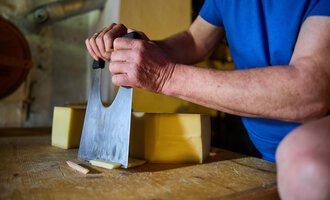 A person is cutting cheese with a large knife on a wooden table. The scene displays craftsmanship and freshness of the ingredients. | © Kleinwalsertal Tourismus eGen - Fotograf: Oliver Farys
