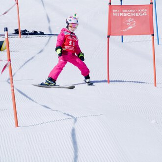 © Skischule Hirschegg | Clemens Paul Ein kleiner Skifahrer in pinker Skiausrüstung fährt durch ein Tor auf einer verschneiten Piste. Im Hintergrund sind weitere Skifahrer und Farbmarkierungen zu sehen. | © Skischule Hirschegg | Clemens Paul