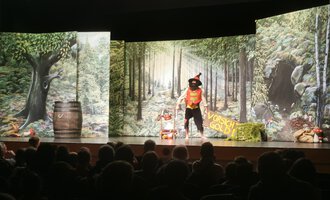 © KiWi Kindertheater GbR A theater performance with a colorful stage design that depicts a forest. An actor in a costume interacts with the audience. | © KiWi Kindertheater GbR