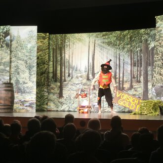 A theater performance with a colorful stage design that depicts a forest. An actor in a costume interacts with the audience. | © KiWi Kindertheater GbR
