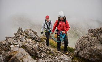 Zwei Bergsteiger bewegen sich an einem felsigen Hang. Es ist neblig und die Umgebung wirkt wild und unberührt. | © Bergschule Kleinwalsertal | Oliver Farys