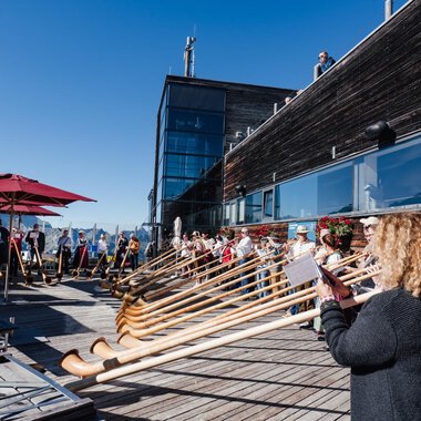 A group of people is playing alphorns on a terrace. In the background, sunny mountains and a clear blue sky can be seen. | © Kleinwalsertal Tourismus | Berthold Schick