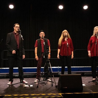 A group of six singers stands on a stage and sings into microphones. All are wearing red and black, and the stage is dimly lit. | © Nice Try