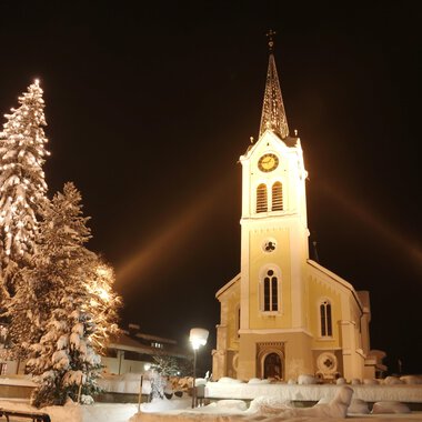 A snowy landscape with a illuminated church and a decorated Christmas tree. The night sky enhances the festive atmosphere. | © Kleinwalsertal Tourismus | Frank Drechsel