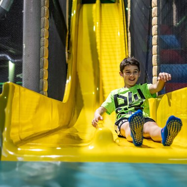 A smiling child happily slides down a yellow slide. In the background, playground equipment can be seen. | © Wonnemar Sonthofen | interSPA Gruppe