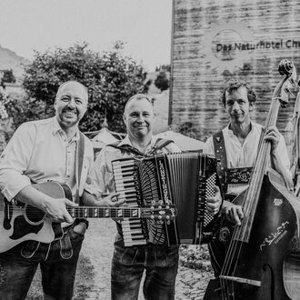 © Naturhotel Chesa Valisa Drei Musiker stehen fröhlich zusammen und spielen verschiedene Instrumente. Im Hintergrund ist das Naturhotel zu sehen. | © Naturhotel Chesa Valisa