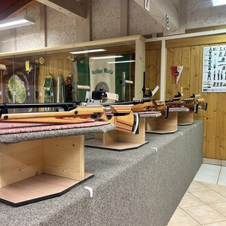 © Luftgewehrstand | Michael Veit Eine Reihe von Sportgewehren auf Ausstellungsständern in einem Schießstand. Im Hintergrund sind Anleitungen und Informationen über den Schießsport zu sehen. | © Luftgewehrstand | Michael Veit