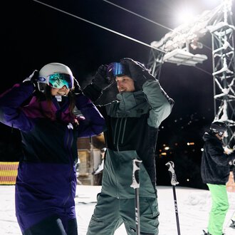 © Kleinwalsertal Tourismus | Oliver Farys A pair of skiers is preparing for night skiing. In the background, other skiers and a lit ski lift can be seen. | © Kleinwalsertal Tourismus | Oliver Farys