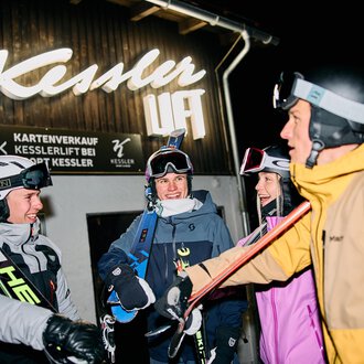 A pair of skiers is preparing for night skiing. In the background, other skiers and a lit ski lift can be seen. | © Kleinwalsertal Tourismus | Oliver Farys