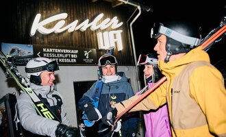 A pair of skiers is preparing for night skiing. In the background, other skiers and a lit ski lift can be seen. | © Kleinwalsertal Tourismus | Oliver Farys